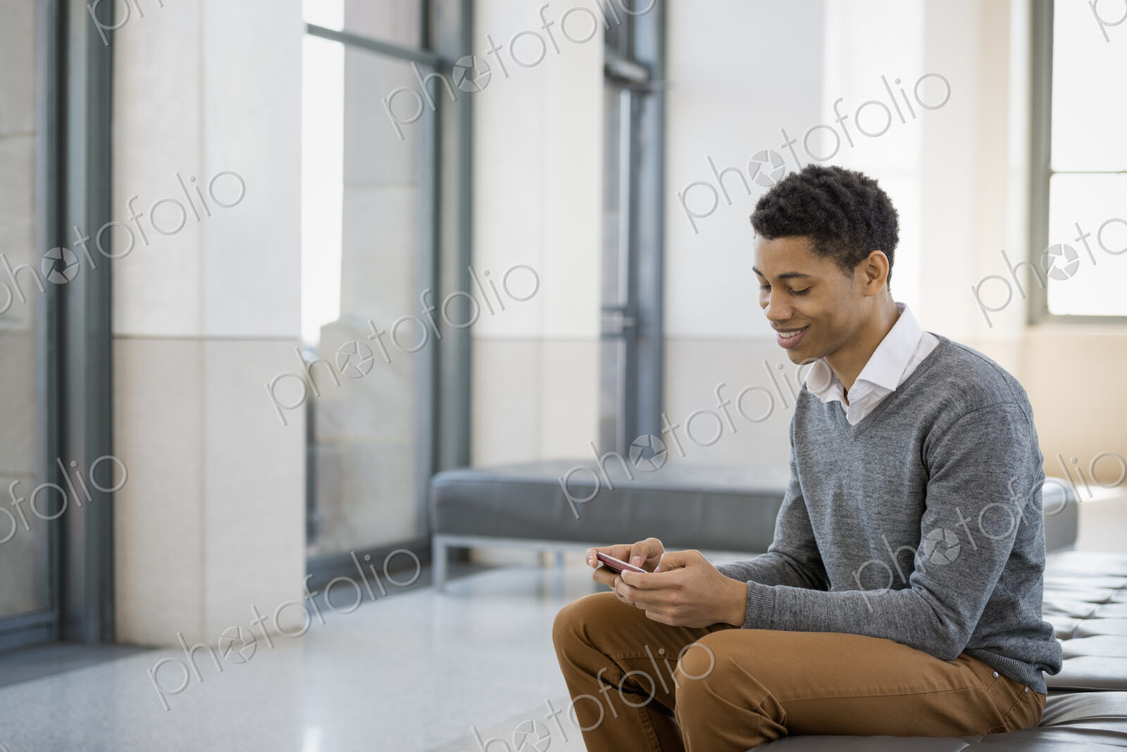 Urban Lifestyle. A young man sitting in a lobby, on a bench seat. Using his mobile phone.