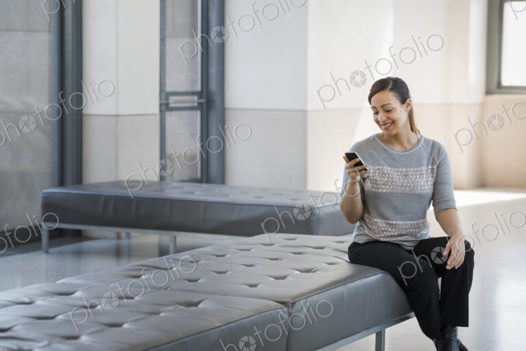 Urban Lifestyle. A young woman sitting on a seat in a building, using her mobile phone.