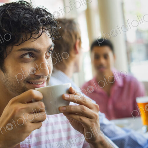 Urban Lifestyle. Three young men around a table in a cafe. One man taking a drink from…