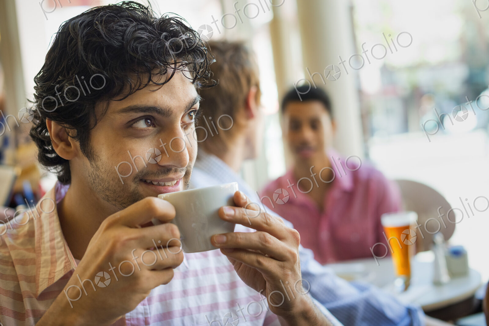 Urban Lifestyle. Three young men around a table in a cafe. One man taking a drink from…