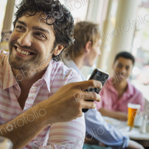 Urban Lifestyle. Three young men around a table in a cafe. One using his smart phone Urban Lifestyle. Three young men around a table in a cafe. One using his smart phone