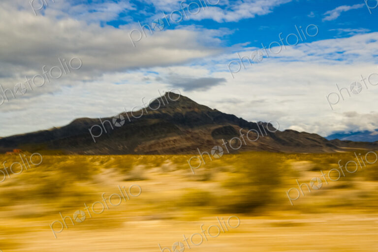 View across a desert landscape to a sharp peak, seen from a moving vehicle.