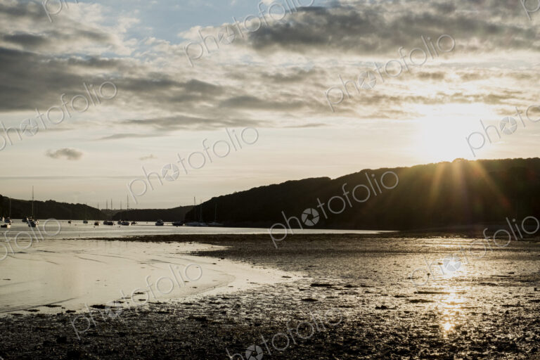 View across a sandy beach towards sailboats and distant cliffs at sunset.