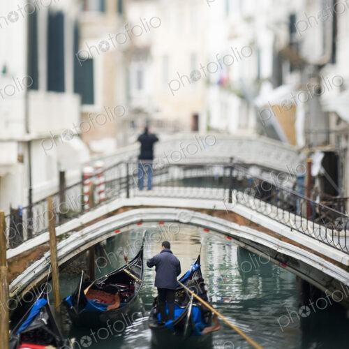 View along a narrow canal, with a person standing on a bridge. A gondola floating below. View along a narrow canal, with a person standing on a bridge. A gondola floating below.