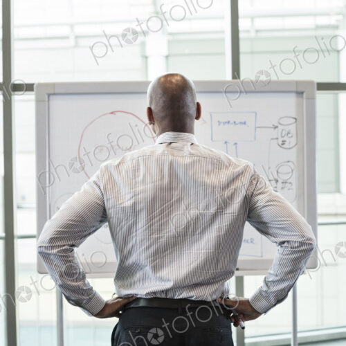 View from behind of a businessman standing at a white board in front of a large window. View from behind of a businessman standing at a white board in front of a large window.