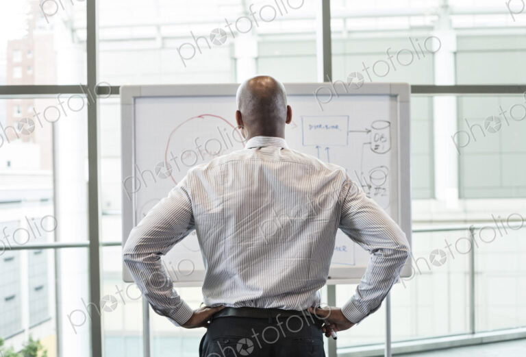 View from behind of a businessman standing at a white board in front of a large window.