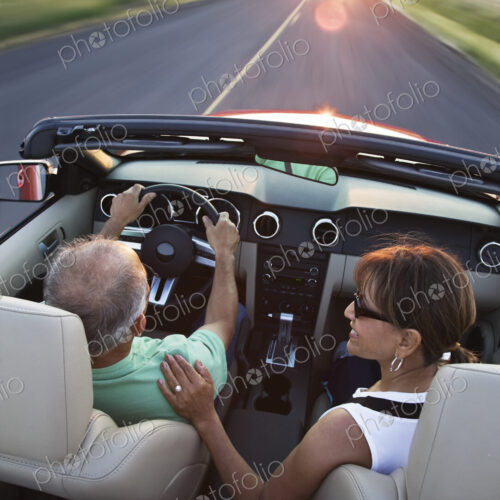 View from behind of a senior couple in a convertible sports car driving on a highway…