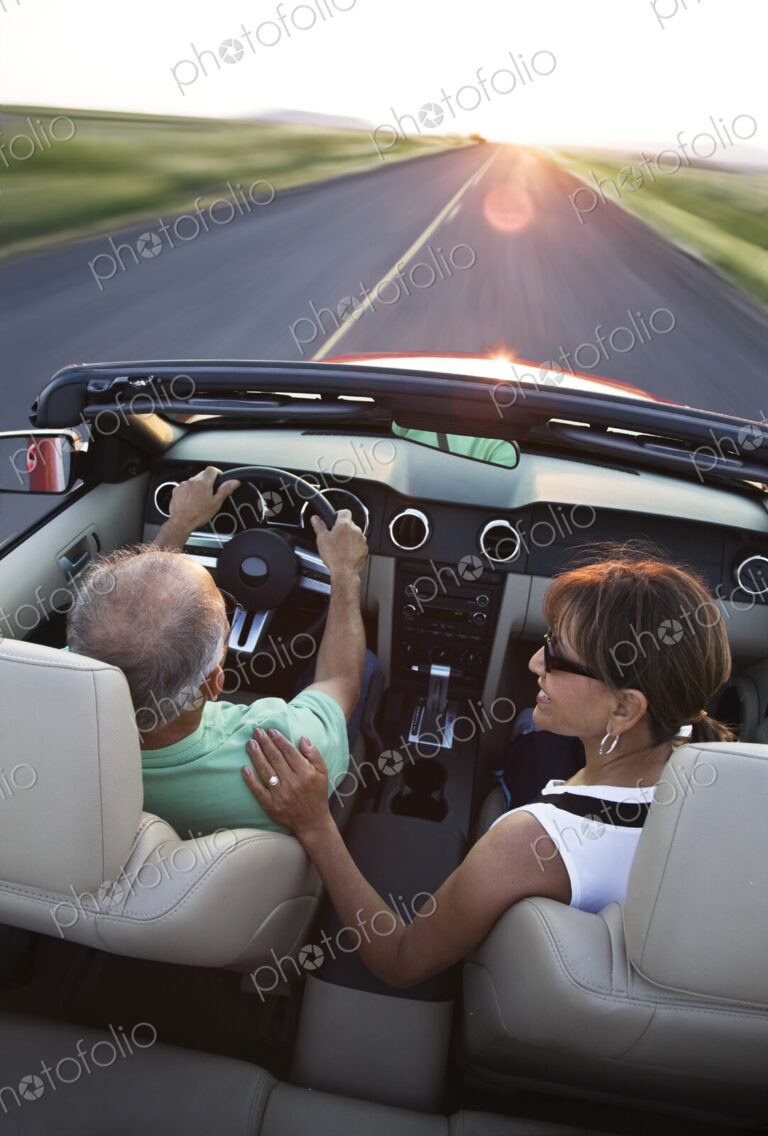 View from behind of a senior couple in a convertible sports car driving on a highway…