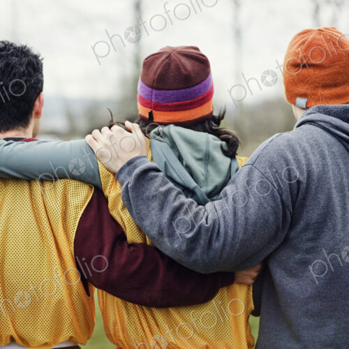 View from behind of three friends with their arms around one another in sports kit and… View from behind of three friends with their arms around one another in sports kit and…