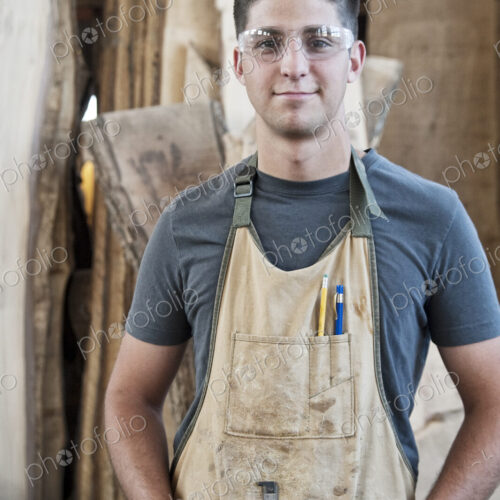 View of a young caucasian man factory worker wearing an apron in a woodworking factory. View of a young caucasian man factory worker wearing an apron in a woodworking factory.