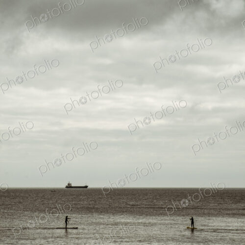 View out to sea from the cliffs, two people paddleboarding on a calm sea, and a large… View out to sea from the cliffs, two people paddleboarding on a calm sea, and a large…