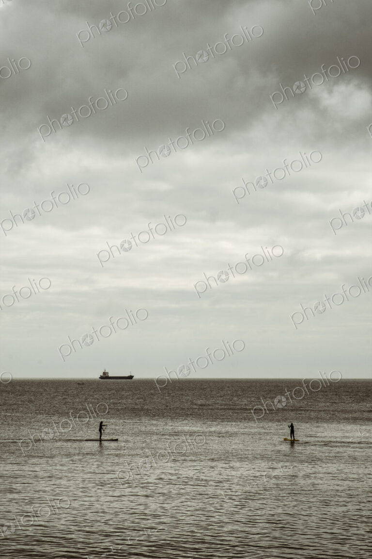 View out to sea from the cliffs, two people paddleboarding on a calm sea, and a large…