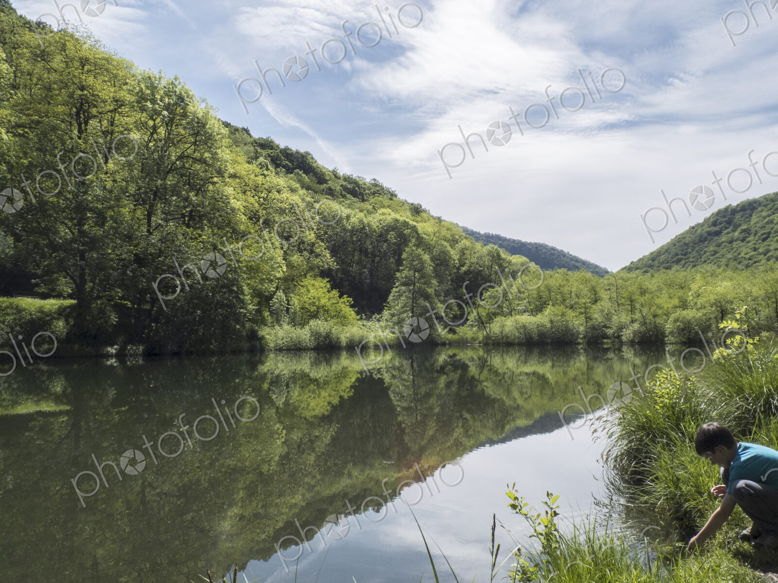 View over flat calm lake water, mountains and woodland, a boy on the shore.