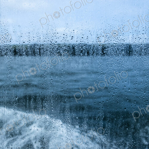 View through a rain spattered window over water and a boat's wake on an overcast day.