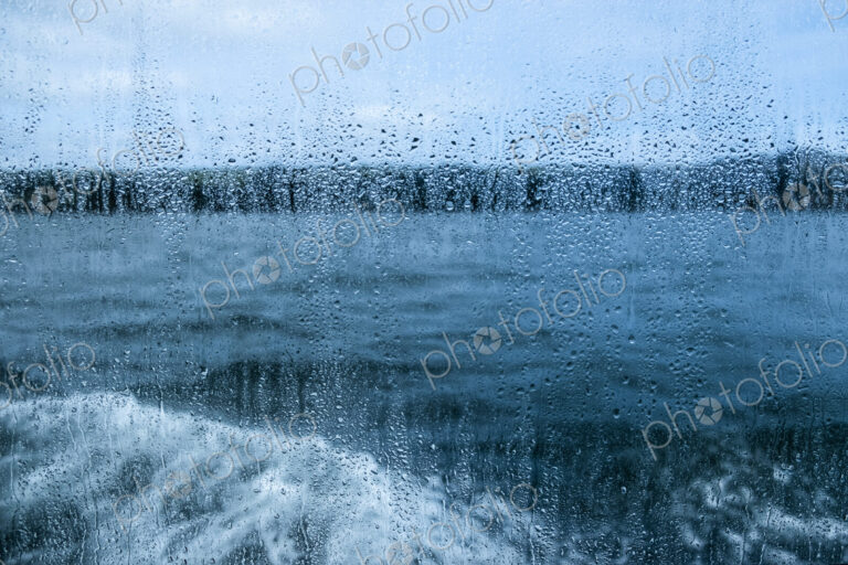 View through a rain spattered window over water and a boat's wake on an overcast day.