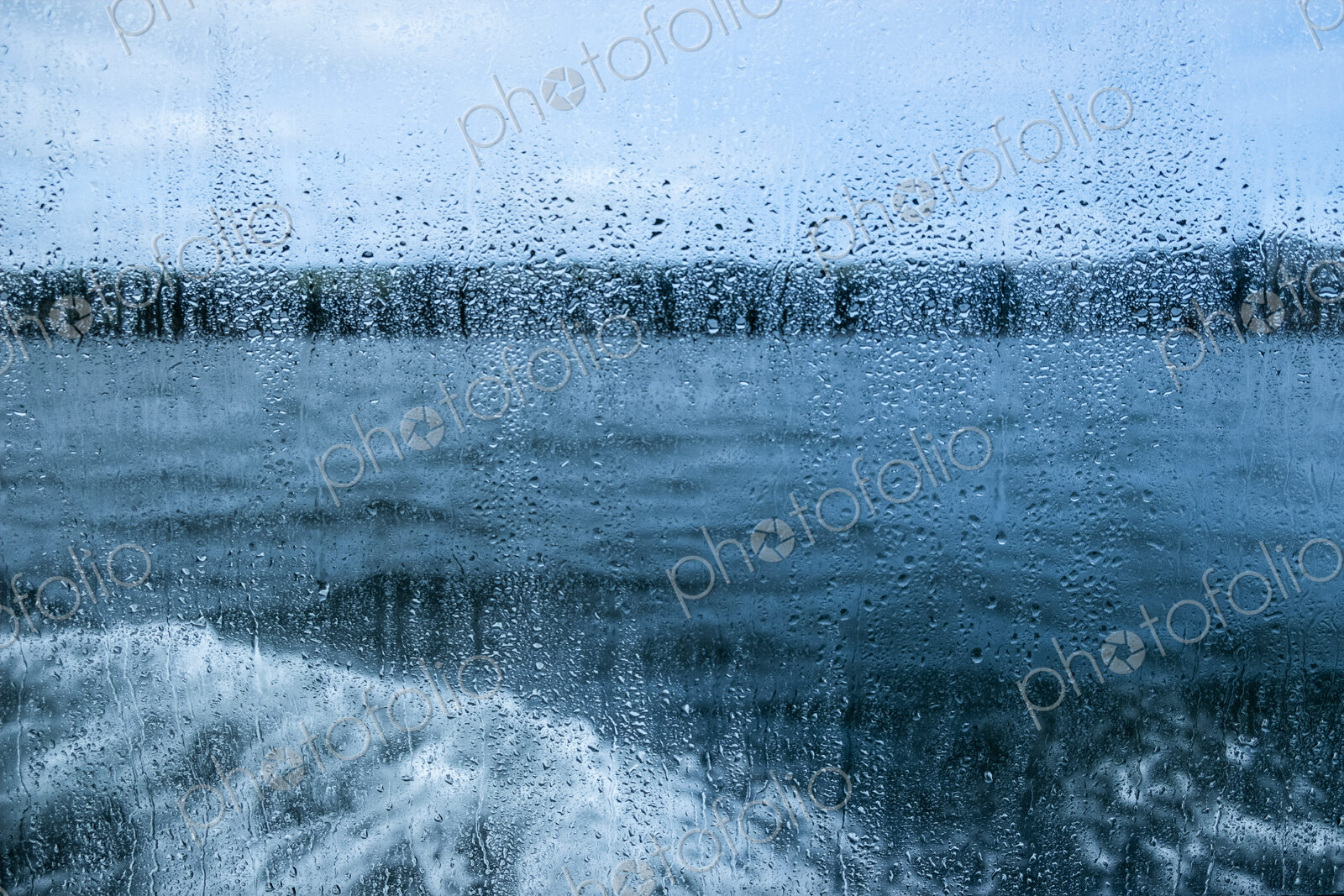 View through a rain spattered window over water and a boat’s wake on an overcast day.