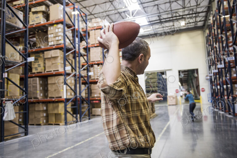 Warehouse employees playing catch with an American football during a work break in a…