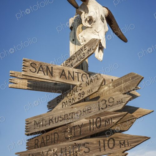 Weathered wooden direction sign topped with a cow skull and horns lists distances to…