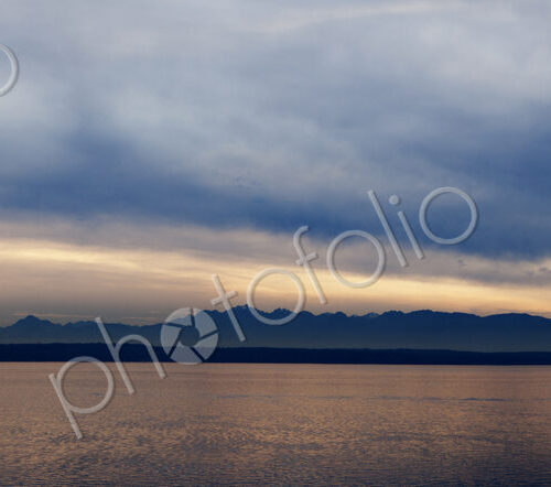 Wide angle view of Puget Sound with Olympic mountain range beyond and cloudy sky.