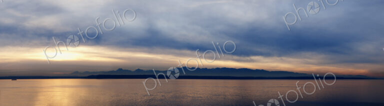 Wide angle view of Puget Sound with Olympic mountain range beyond and cloudy sky.