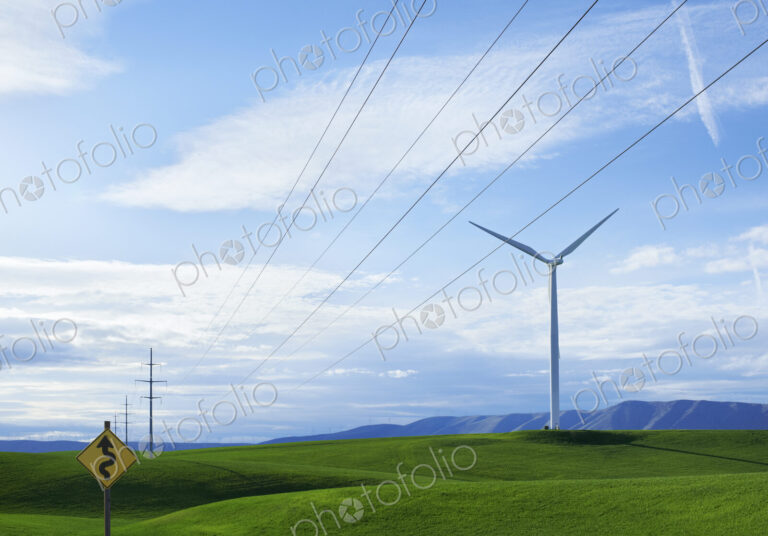 Wind turbines on a grassy hilltop, and a road sign warning of a Winding Road. Green…