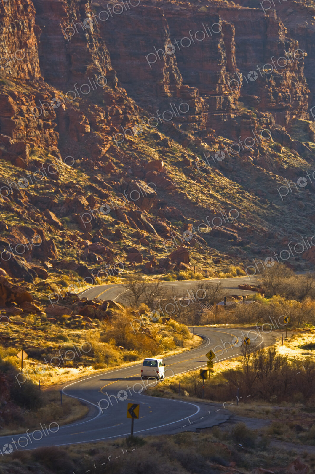 Winding road with a single car, dramatic sunlit cliffs rising from a valley