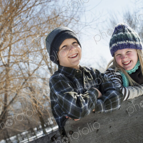 Winter scenery with snow on the ground. Two children in knitted hats leaning on a fence.