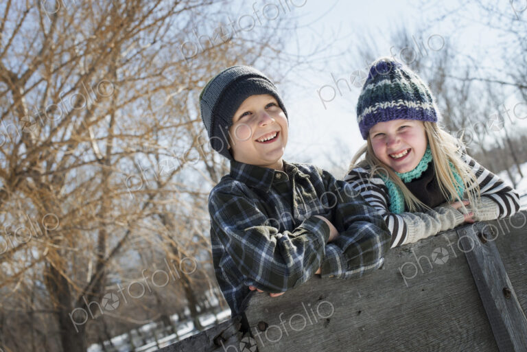 Winter scenery with snow on the ground. Two children in knitted hats leaning on a fence.