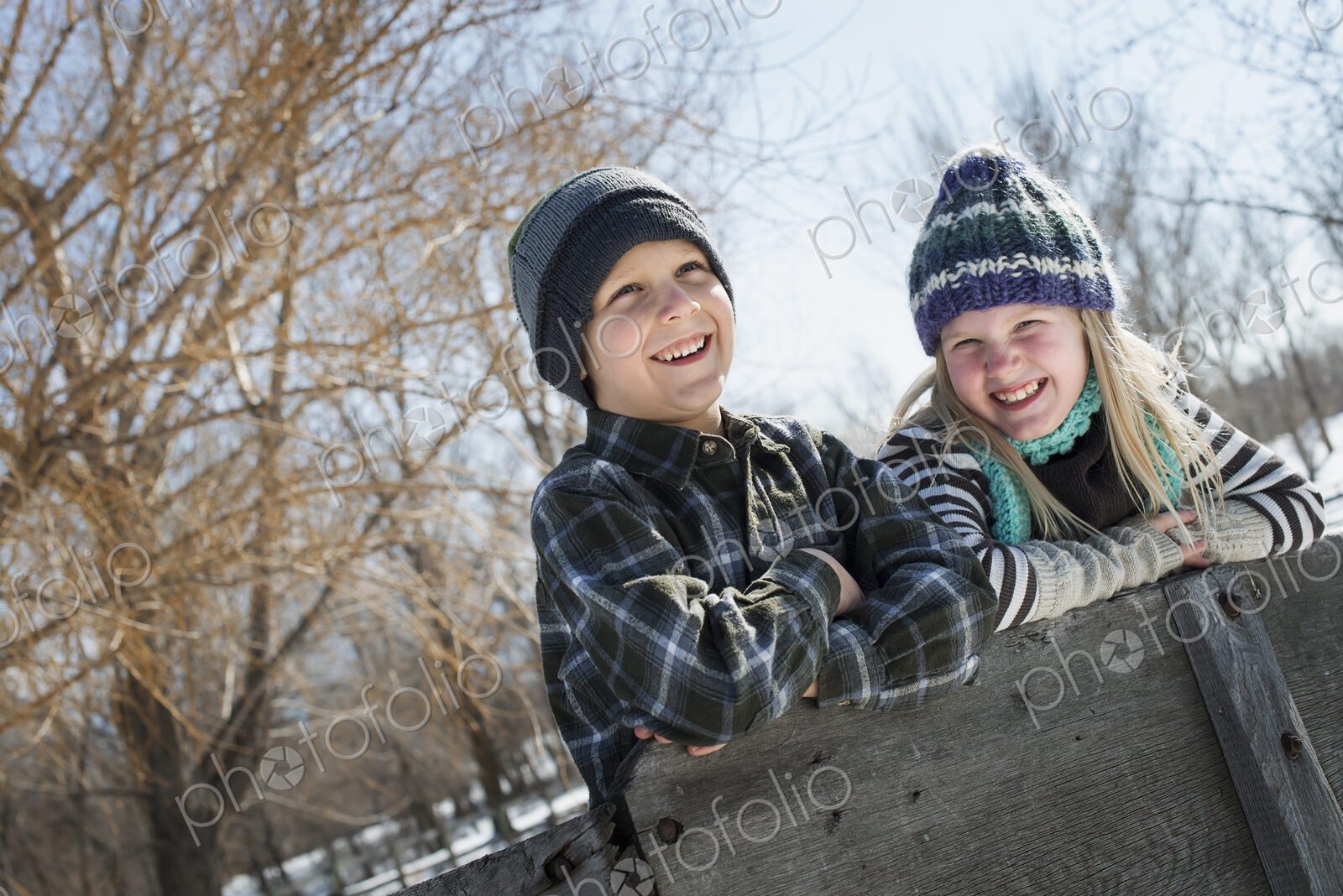 Winter scenery with snow on the ground. Two children in knitted hats leaning on a fence.