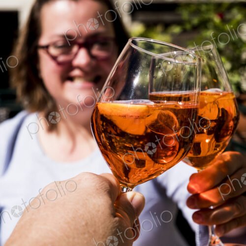 Woman and another person sitting at a table toasting with wine glasses of Aperol Spritz.