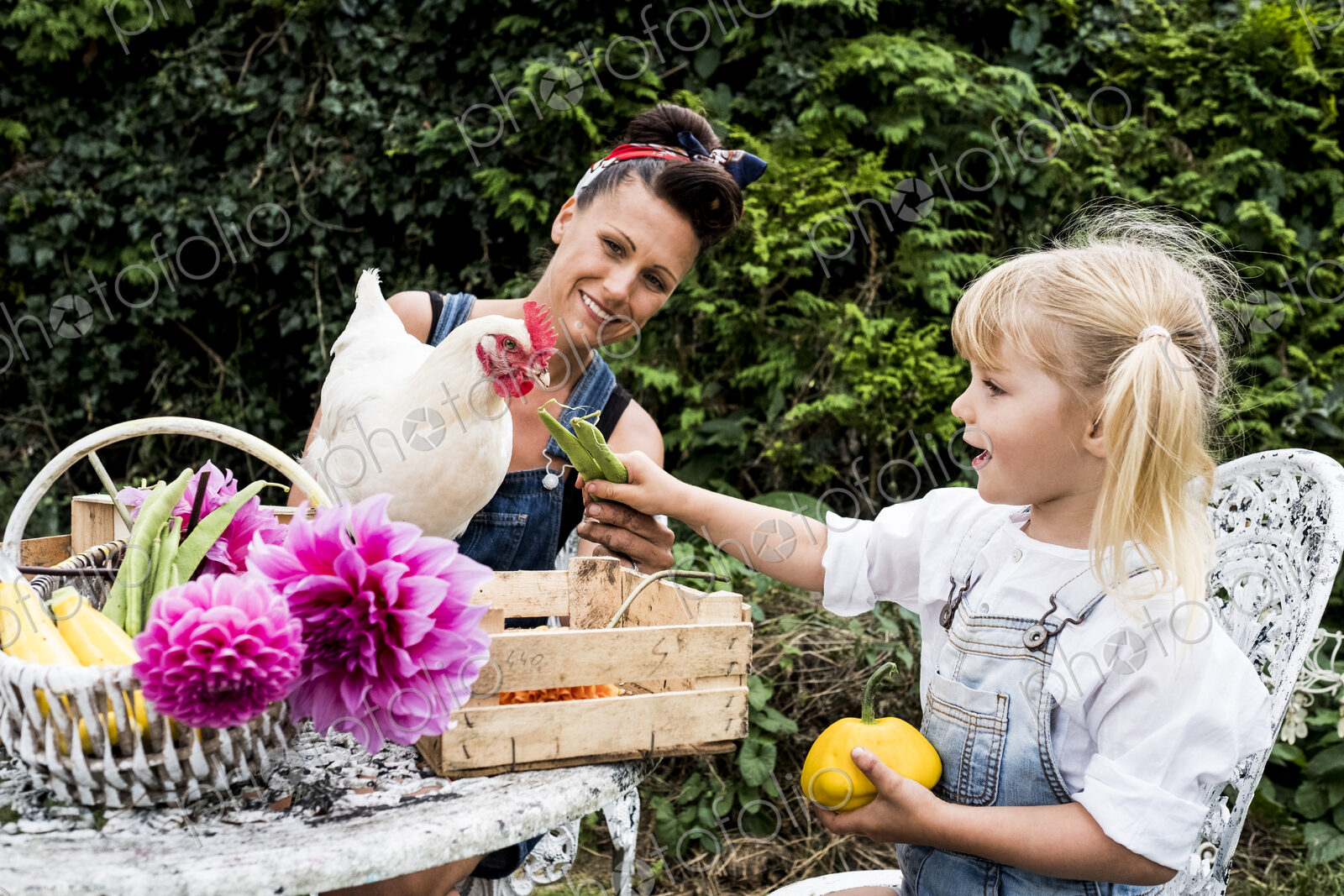 Woman and girl sitting at a table in a garden, feeding white chicken.