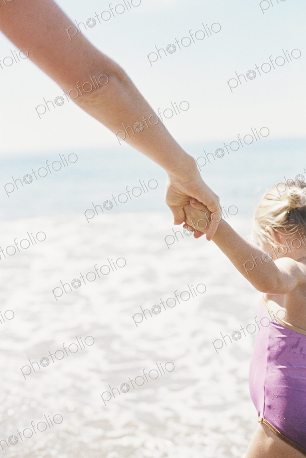 Woman and her daughter holding hands, standing on a sandy beach by he ocean.
