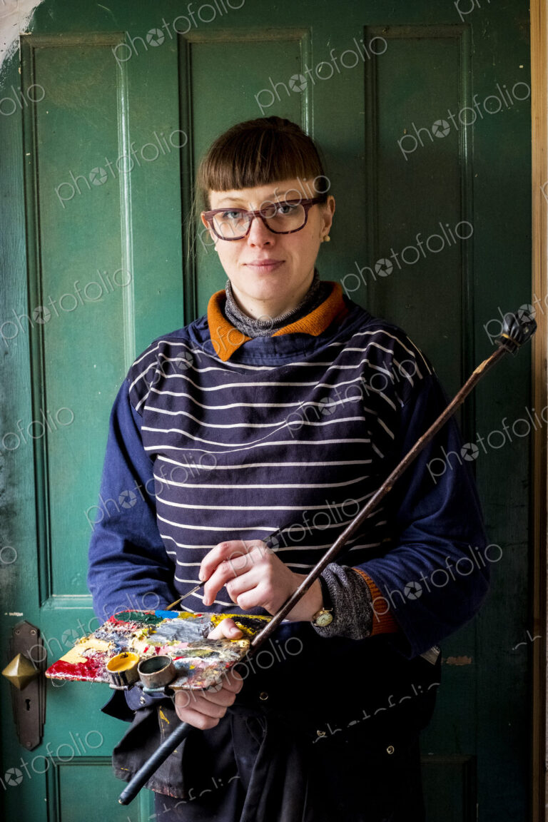 Woman artist wearing glasses holding paintbrush, palette and Mahlstick, looking at camera.