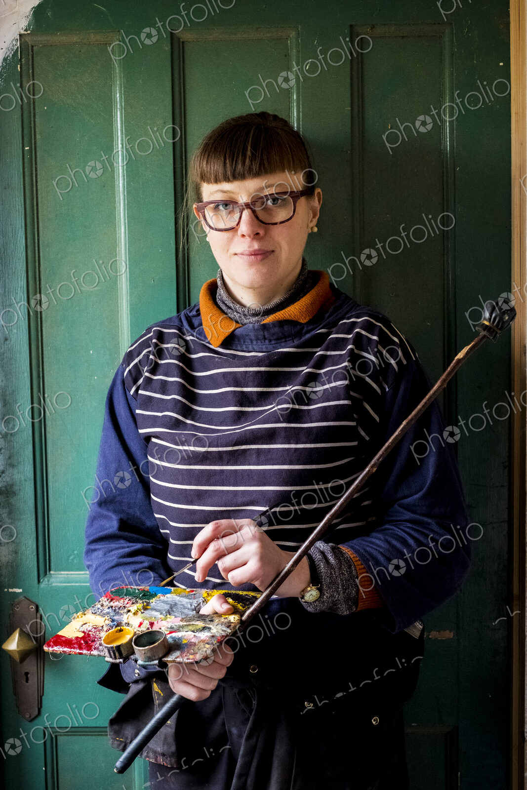 Woman artist wearing glasses holding paintbrush, palette and Mahlstick, looking at camera.