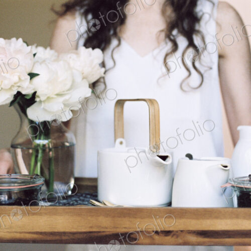 Woman carrying a tray with a teapot and a vase of white roses.