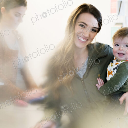 Woman holding a baby standing next to a cash register with shop assistant, smiling at…