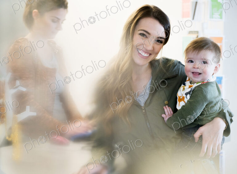 Woman holding a baby standing next to a cash register with shop assistant, smiling at…