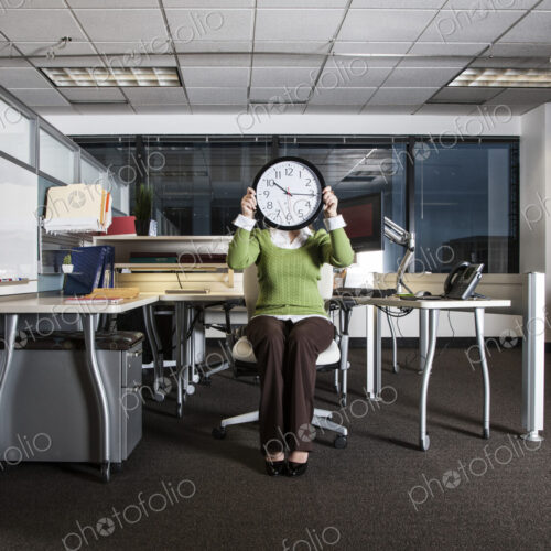 Woman holding a clock face while sitting in her cubicle office space. Woman holding a clock face while sitting in her cubicle office space.