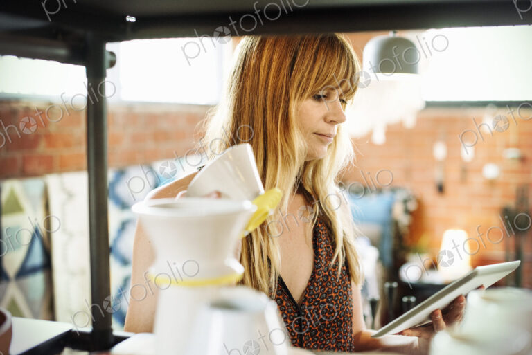 Woman in a shop, holding a digital tablet and small ceramic pot.