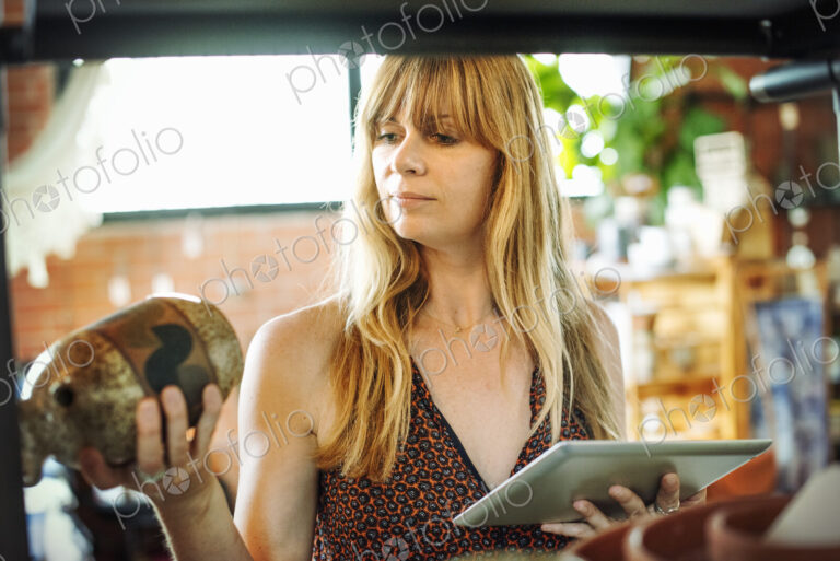 Woman in a shop, holding a digital tablet and small ceramic vase.