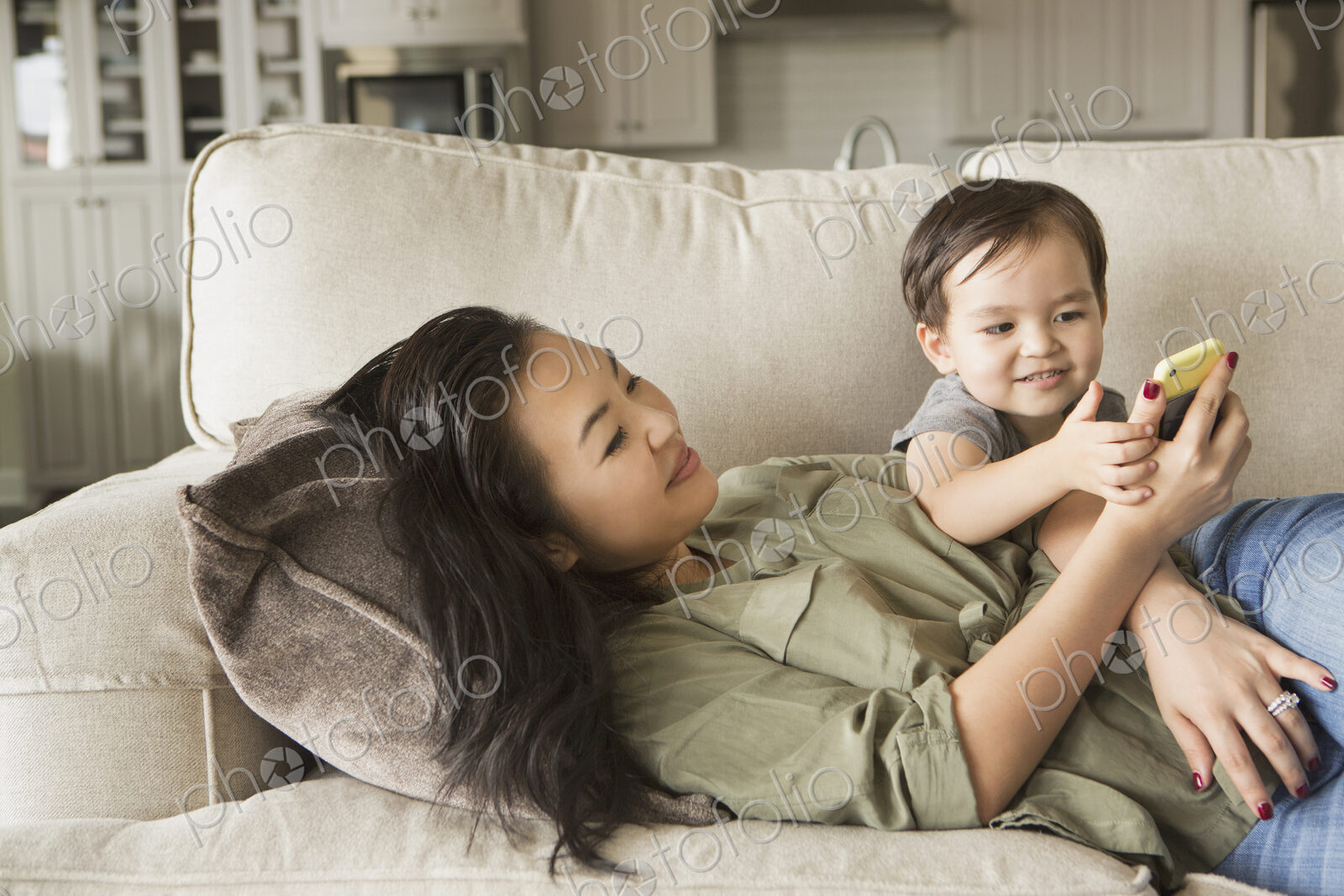 Woman lying on a sofa, smiling, cuddling with her young son and looking at a cell phone.