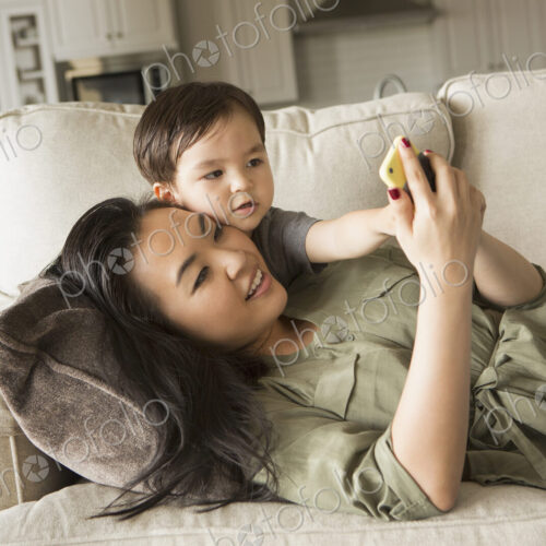 Woman lying on a sofa, smiling, cuddling with her young son and looking at a cell phone. Woman lying on a sofa, smiling, cuddling with her young son and looking at a cell phone.
