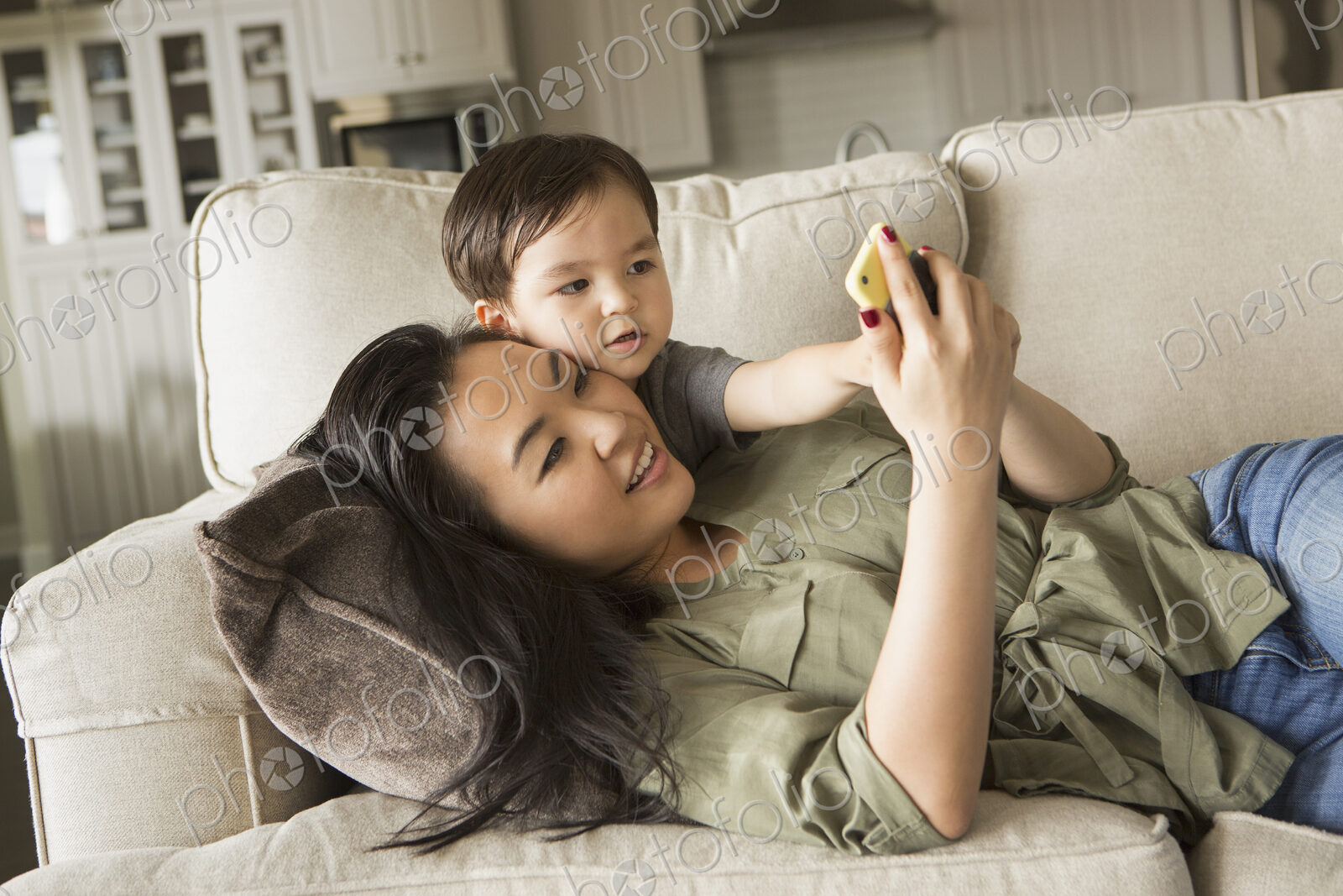 Woman lying on a sofa, smiling, cuddling with her young son and looking at a cell phone.