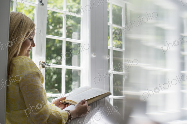 Woman sitting by a window, reading a book.