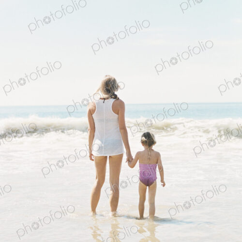Woman standing hand in hand with her daughter on a sandy beach.