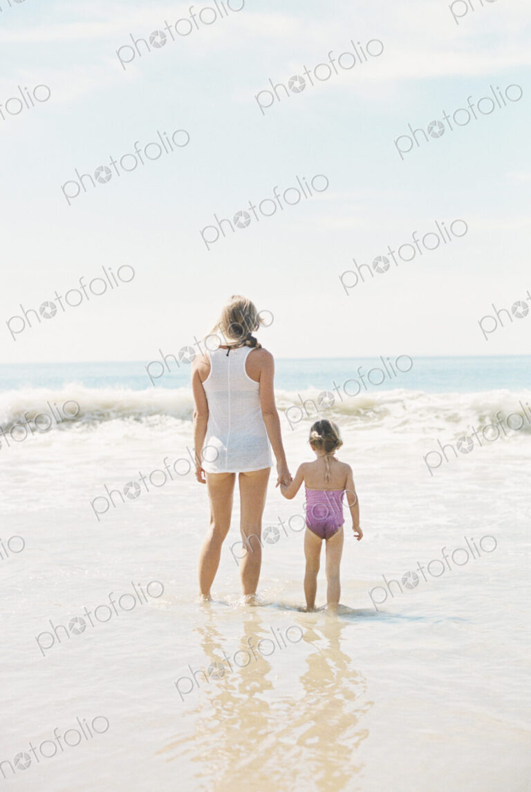 Woman standing hand in hand with her daughter on a sandy beach.
