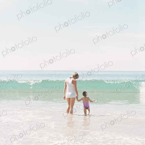 Woman standing hand in hand with her daughter on a sandy beach watching a wave.
