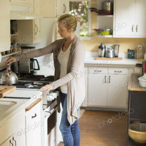 Woman standing in a kitchen, placing a kettle on a stove.