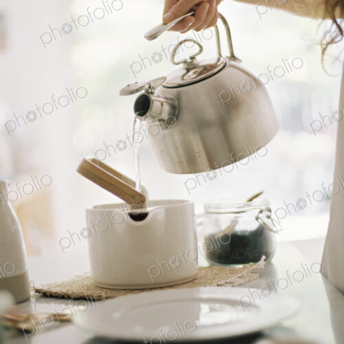 Woman standing in a kitchen pouring hot water from a kettle into a tea pot.