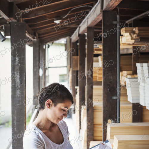 Woman standing in a lumber yard, holding a folder, checking notes.